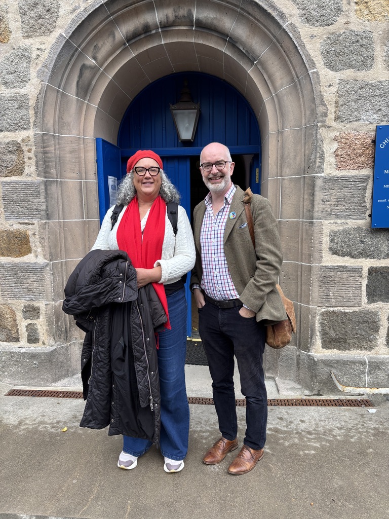 The photo shows two people standing in front of a stone building with a blue arched door. The person on the left is wearing a red hat, red scarf, white top, and blue jeans, and is holding a black coat. The person on the right is wearing a brown jacket, checked shirt, and dark trousers with brown shoes. Both are smiling. The photo shows two people standing in front of a stone building with a blue arched door. The person on the left is wearing a red hat, red scarf, white top, and blue jeans, and is holding a black coat. The person on the right is wearing a brown jacket, checked shirt, and dark trousers with brown shoes. Both are smiling.